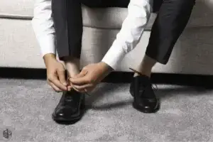 square_4_media A person in dress pants and a white shirt sits on a couch, tying the laces of black dress shoes on a grey carpet. media_agency