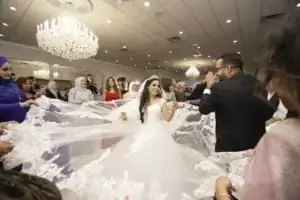 square_4_media A bride in a white gown and veil smiles while surrounded by guests holding her long lace train at a wedding reception in a decorated hall. media_agency