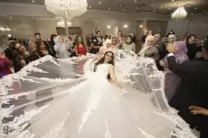 square_4_media A bride in a white wedding dress with a long, lace-trimmed train is surrounded by guests celebrating in a decorated ballroom. media_agency