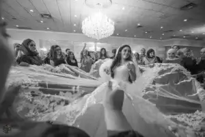 square_4_media A bride in a wedding dress stands smiling as guests surround her, holding up her long, lace-trimmed veil in a decorated reception hall. media_agency