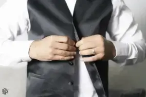 square_4_media A person wearing a white shirt and black vest is fastening the buttons on the vest. The person has a ring on their left hand. media_agency