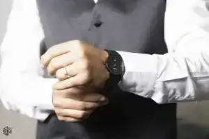 square_4_media A person in a dress shirt and vest adjusts a black wristwatch on their left wrist; a silver ring is visible on their ring finger. media_agency