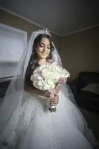 square_4_media A bride in a white wedding dress and veil holds a bouquet of white and pink flowers, standing indoors and smiling. media_agency