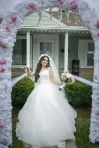 square_4_media A bride in a white wedding dress and veil stands under a floral arch, holding a bouquet, with a house and greenery in the background. media_agency