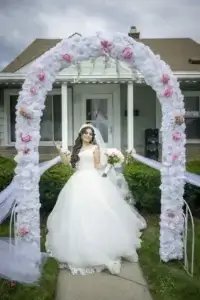 square_4_media A bride in a white wedding dress stands under a floral archway in front of a house, holding a bouquet and smiling at the camera. media_agency