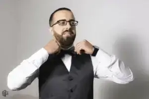 square_4_media A man wearing glasses adjusts his black bow tie and vest over a white shirt, standing against a plain light background. media_agency