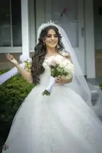 square_4_media A bride in a white wedding dress and veil stands outside, holding a bouquet of white and pink roses, smiling. media_agency