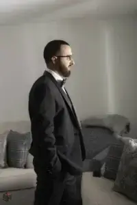 square_4_media A man in a black suit and glasses stands in profile in a living room, hands in pockets, next to a light-colored sofa with patterned cushions. media_agency