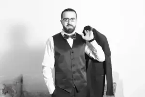 square_4_media A man in a formal vest, bow tie, and glasses stands indoors holding a suit jacket over his shoulder, with one hand in his pocket. media_agency