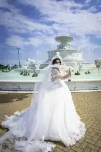 square_4_media A bride in a white wedding dress stands in front of a large decorative fountain under a partly cloudy sky, holding her veil and bouquet. media_agency