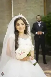 square_4_media A bride in a white dress and veil holds a bouquet of roses and smiles at the camera; a groom stands blurred in the background. media_agency