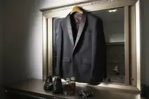 square_4_media A black suit jacket on a hanger is displayed on a mirror above a dresser with black dress shoes, cologne bottles, and a bow tie neatly arranged. media_agency