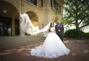square_4_media A bride in a white gown and groom in a dark suit pose outdoors in front of a stone building; the bride’s veil is flowing in the wind. media_agency