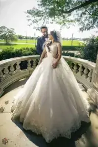 square_4_media A bride in a white gown and a groom in a suit stand together on a stone balcony, surrounded by greenery and sunlight. media_agency