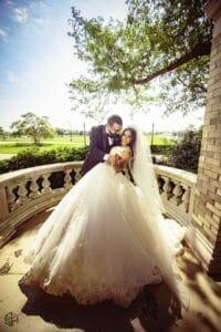 square_4_media A bride in a white gown and a groom in a black suit embrace on a stone terrace with trees and green landscape in the background. media_agency