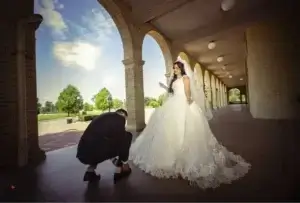 square_4_media A groom crouches to adjust the bride’s gown under an arched walkway, while the bride stands smiling in a white wedding dress and veil. media_agency
