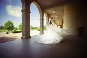 square_4_media A woman in a white wedding dress stands under an arched walkway, her dress fanned out as if she is spinning, with trees and blue sky visible in the background. media_agency