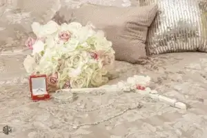 square_4_media A bouquet of white and pink roses, a red ring box with a ring, bracelets, jewelry, and accessories arranged on a beige patterned bedspread with decorative pillows. media_agency