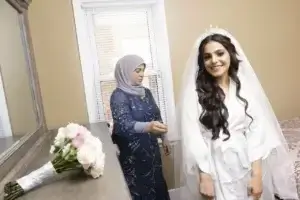square_4_media A bride in a white robe and veil stands smiling in a bedroom while a woman in a blue dress and hijab stands nearby. A bouquet of flowers rests on the dresser. media_agency