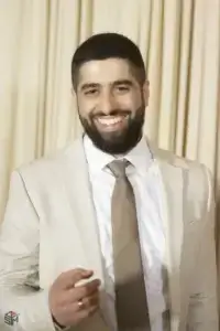 square_4_media A man wearing a light beige suit and brown tie is smiling at the camera, standing in front of cream-colored curtains. media_agency