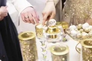 square_4_media Two people in formal attire touch a decorative gold cup surrounded by gold and white sweets and ornate tableware at an event or celebration. media_agency