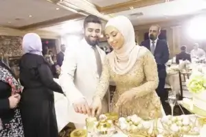 square_4_media A man and a woman dressed in formal attire smile as they cut a dessert together at a decorated indoor event, with guests and tables in the background. media_agency