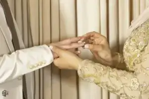 square_4_media A person in a beige and gold outfit is placing a ring on another person's finger, both standing in front of cream-colored curtains. media_agency