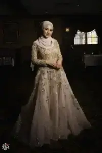 square_4_media A woman wearing a light pink hijab and an embroidered, gold-accented gown stands smiling in a dimly lit room with carpeted floors and framed art on the walls. media_agency
