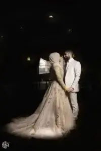 square_4_media A couple dressed in formal attire, with the woman in an embellished gown and hijab and the man in a light-colored suit, stand facing each other holding hands in a dimly lit room. media_agency