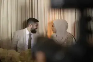 square_4_media A man in a suit and a woman in a hijab sit next to each other, smiling and making eye contact, with curtains and floral arrangements in the background. media_agency