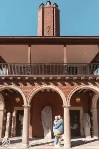 square_4_media Two people stand in front of a brick building with arches and a prominent chimney on a clear day. media_agency