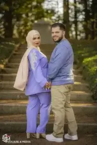 square_4_media A woman in a lavender outfit and hijab stands holding hands with a man in a blue sweater and khaki pants on outdoor steps, facing the camera. media_agency