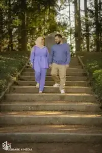 square_4_media A woman in a light purple outfit and hijab walks down outdoor steps holding hands with a man in a blue sweater and beige pants. Trees and sunlight are visible in the background. media_agency