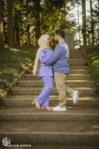 square_4_media A couple stands facing each other on outdoor steps surrounded by greenery, with the man lifting one leg backward and both smiling. media_agency