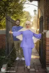 square_4_media A woman in a purple outfit and hijab stands holding an arched gate, while a man in a blue shirt stands behind her on a brick pathway outdoors. media_agency