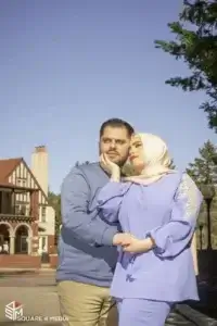 square_4_media A man and woman, both wearing blue outfits, stand closely together outdoors near a building with Tudor-style architecture under a clear blue sky. media_agency