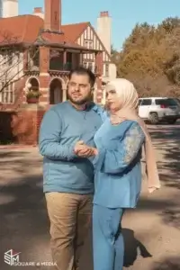 square_4_media A man and woman stand holding hands on a sunny street in front of a large brick house. The woman wears a light pink hijab and blue outfit; the man wears a blue sweater and beige pants. media_agency