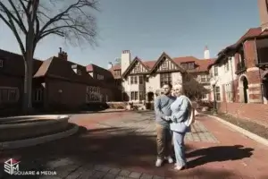 square_4_media A man and woman stand together in front of a large, historic brick building with gabled roofs and multiple chimneys on a sunny day. media_agency