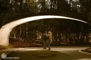 square_4_media A couple stands facing each other under a curved concrete bridge in a wooded park area, with sunlight filtering through the trees. media_agency