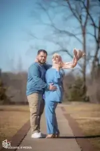 square_4_media A man and woman stand on a walkway outdoors; the woman wears a hijab and blue outfit, while the man's arm is around her waist. The background shows bare trees and a clear sky. media_agency