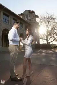 square_4_media A couple stands facing each other and holding hands outside a large building, with sunlight streaming through a tree in the background. media_agency