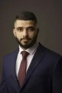 square_4_media A man with short hair and a beard wears a dark blue suit, white shirt, and maroon tie, posing against a plain dark background. media_agency