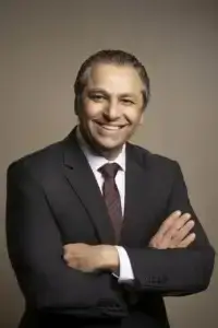 square_4_media Man in a dark suit and red striped tie stands with arms crossed, smiling at the camera against a plain beige background. media_agency