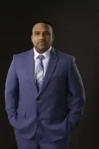 square_4_media A man in a blue suit, white shirt, and striped tie stands with hands in his pockets against a plain dark background. media_agency