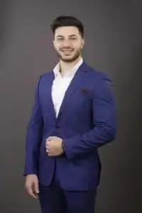 square_4_media A man in a blue suit and white shirt stands against a plain gray background, smiling with one hand holding his jacket. media_agency