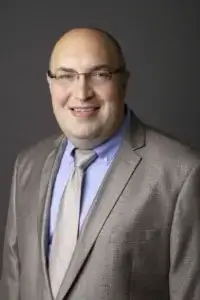 square_4_media A man wearing glasses, a tan suit, light blue shirt, and beige tie, smiling at the camera against a plain dark background. media_agency