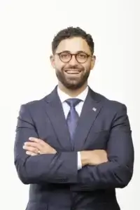 square_4_media A man in a navy suit and tie stands with arms crossed, smiling at the camera against a plain white background. media_agency