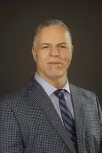 square_4_media A middle-aged man with short gray hair and a goatee wearing a blue checked suit jacket, striped tie, and collared shirt poses against a plain dark background. media_agency