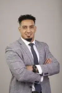 square_4_media A man in a checked grey suit, white shirt, and navy tie stands with arms crossed, smiling against a plain light background. media_agency