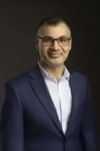 square_4_media A man with short hair, glasses, and a trimmed beard is wearing a blue suit jacket over a white shirt, posing against a dark background. media_agency